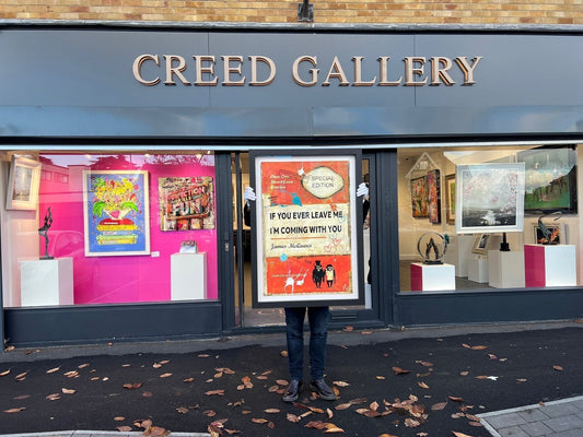 A person in white gloves holding a large, framed James McQueen original titled "If You Ever Leave Me I’m Coming With You" in front of the Creed Gallery storefront in Ascot.