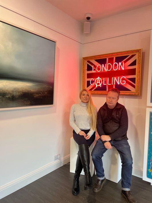 Helena and artist Mark Sloper seated together beneath one of his neon artworks at Creed Gallery