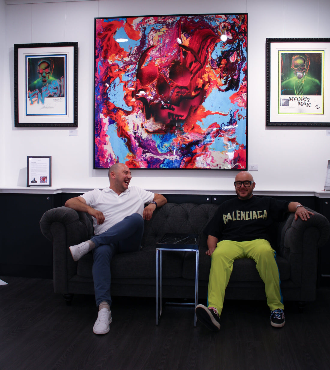 Interior of Creed Gallery featuring Lincoln Townley's 'Success' collection. A large, vibrant pink and blue skull painting hangs above two men on a Chesterfield sofa, flanked by 'Energy' and 'Money Man' prints.