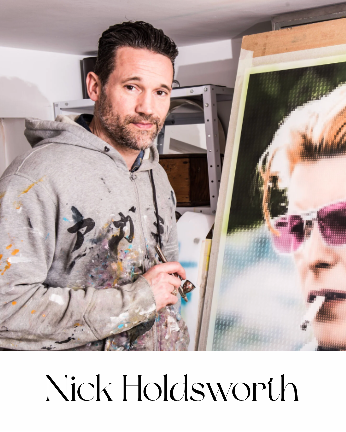 Nick Holdsworth photographed in his studio beside a pixelated portrait work in progress, wearing a paint-splattered hoodie