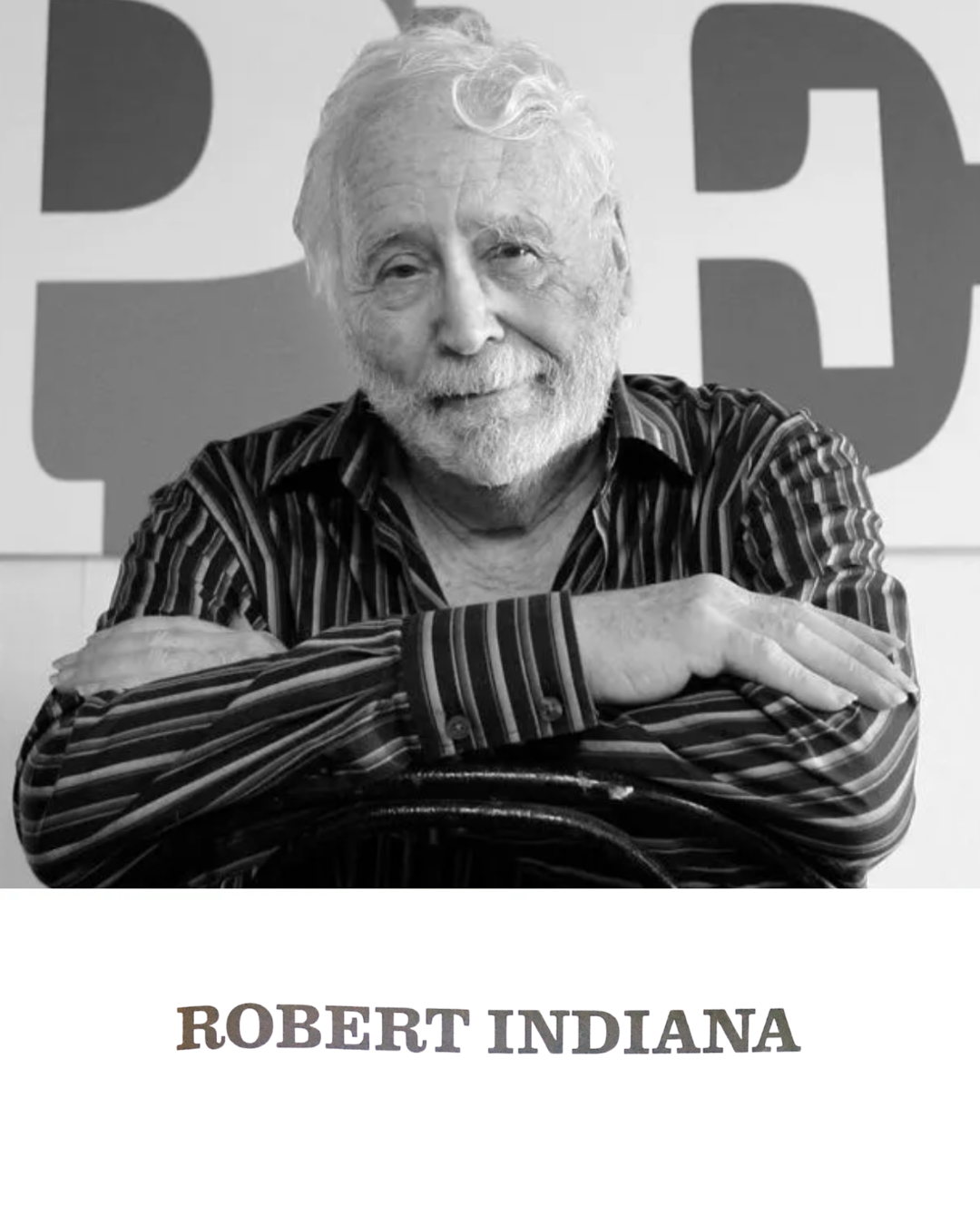 Black-and-white portrait of Pop artist Robert Indiana, seated with arms crossed in front of a large typographic sculpture