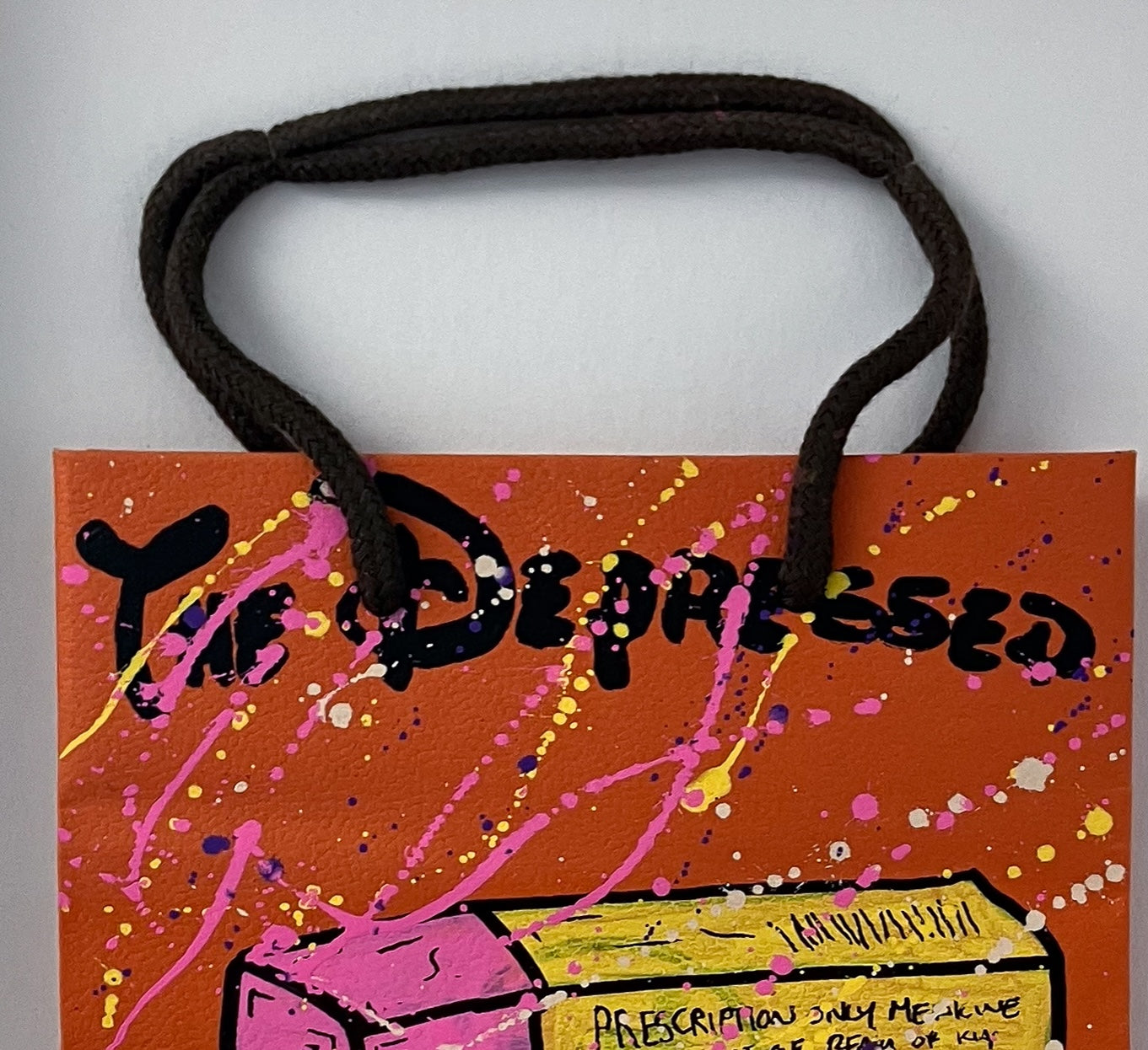 A side-angle view of the "The Depressed Generation" Hermes edition, showing the texture of the branded paper bag and pink spray-paint splatters. The image highlights the 24 x 37.5 cm white box frame and the unique mounting of the contemporary mixed-media work.